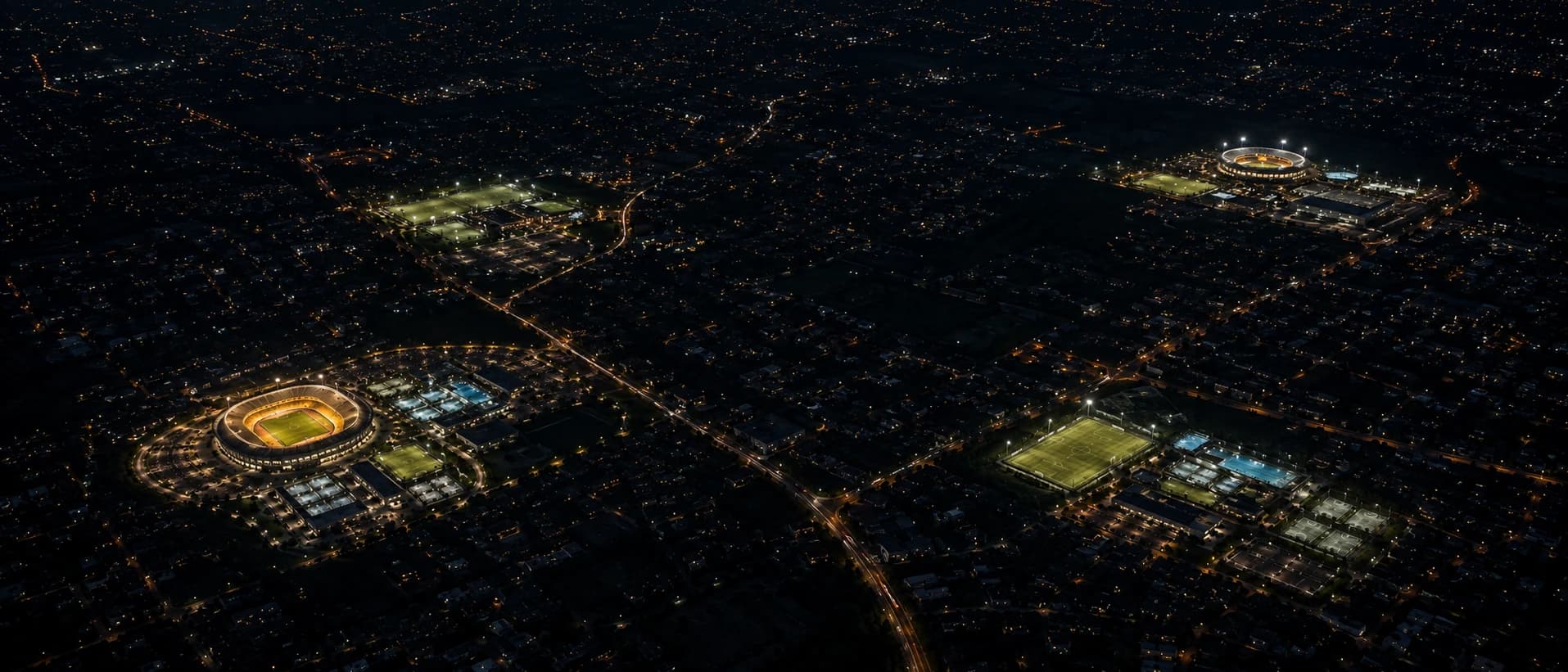 Night aerial of a city with four separate sports facilities lit up across it — each a stadium, pool or court cluster of its own