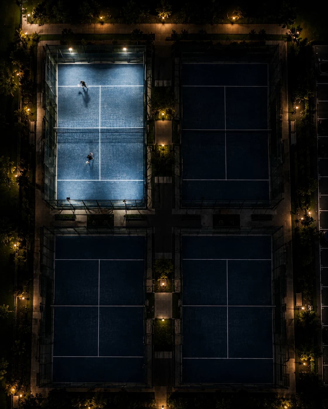 Four padel courts seen from directly above at night — one lit with two players mid-rally, the other three dark but fully marked out